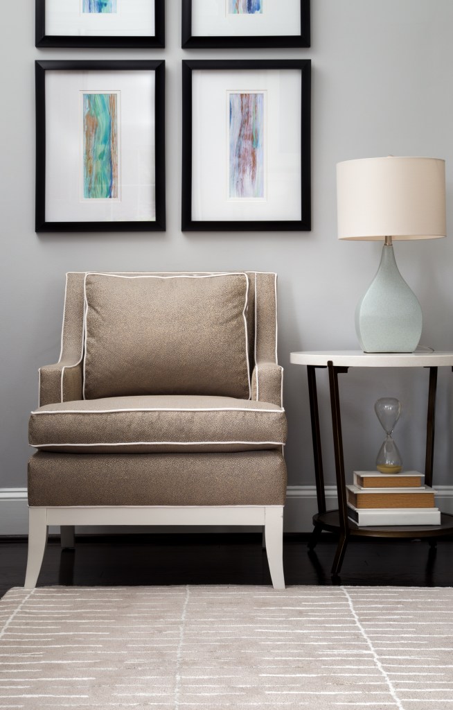 Stylish beige armchair with ornate piping, contemporary side table with books, abstract wall art.