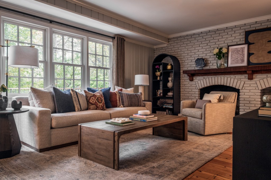 Bright living space featuring a beige sofa, armchair, brick fireplace, and large windows with view of greenery, tasteful interior design.