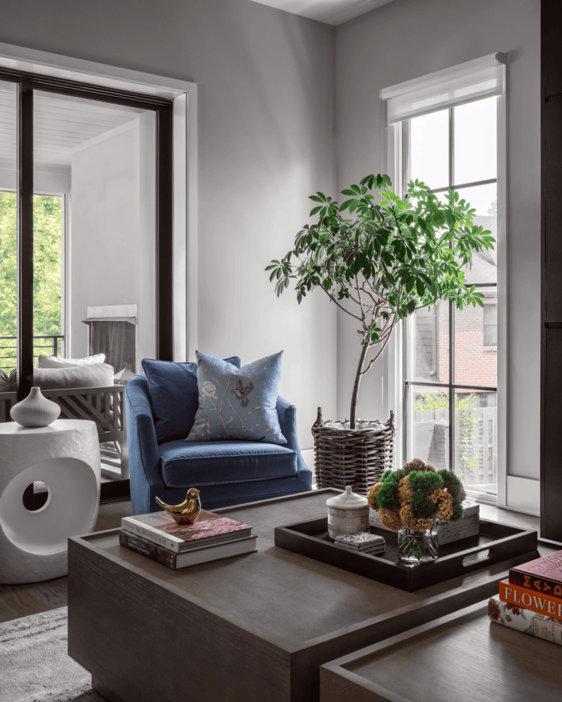 A modern living room featuring a blue armchair with decorative pillows, a wooden coffee table with assorted items, and a potted plant next to large windows that allow natural light to fill the space.