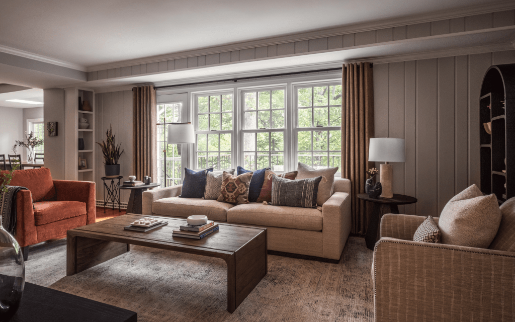 Cozy living room with a beige sofa and colorful pillows, an orange armchair, a wooden coffee table, and large windows with natural light.