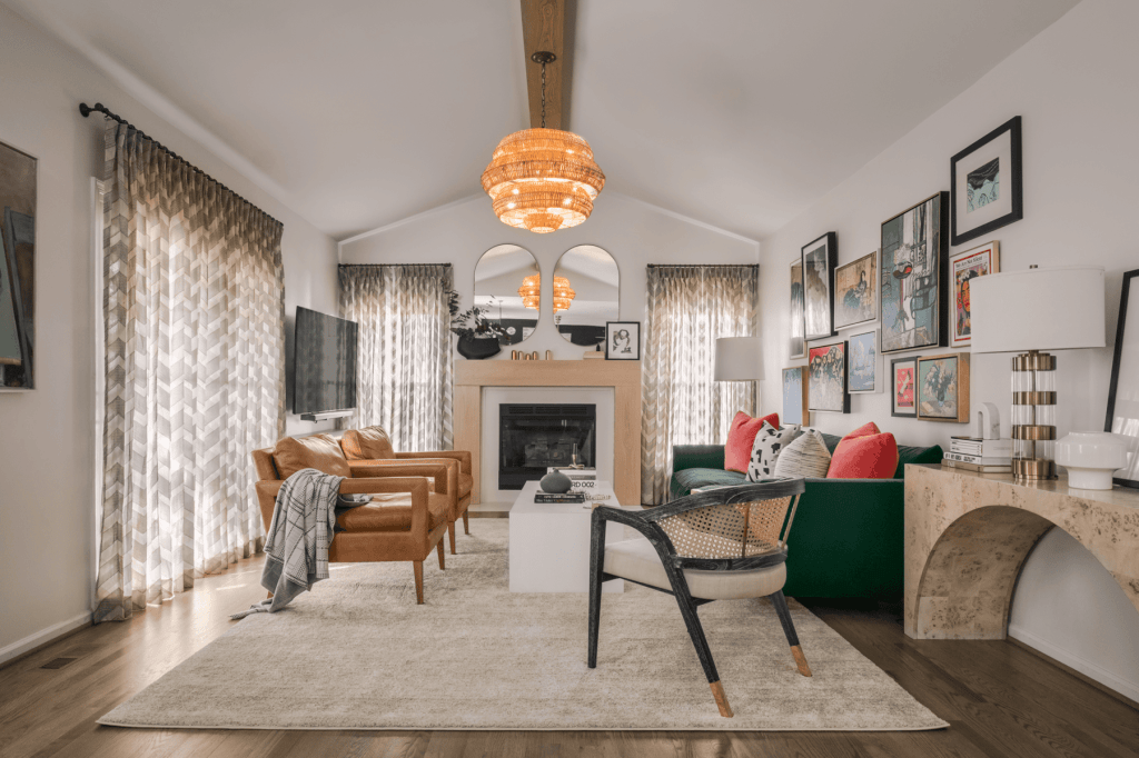 A stylish living room featuring two brown leather chairs, a green couch with red and blue pillows, a white coffee table, and a modern chandelier. The room has large patterned curtains and a fireplace with artwork on the walls.