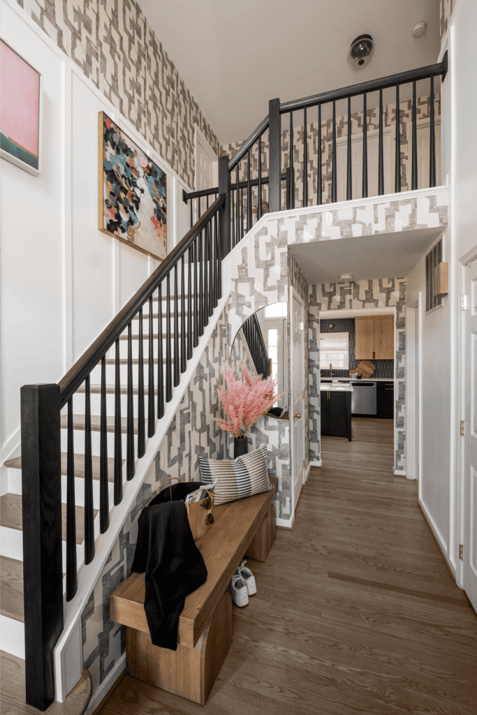 A stylish entryway featuring a staircase with black railings, a wooden bench with cushions, and decorative art on the walls. The wallpaper displays a geometric pattern, complemented by plants and modern decor.