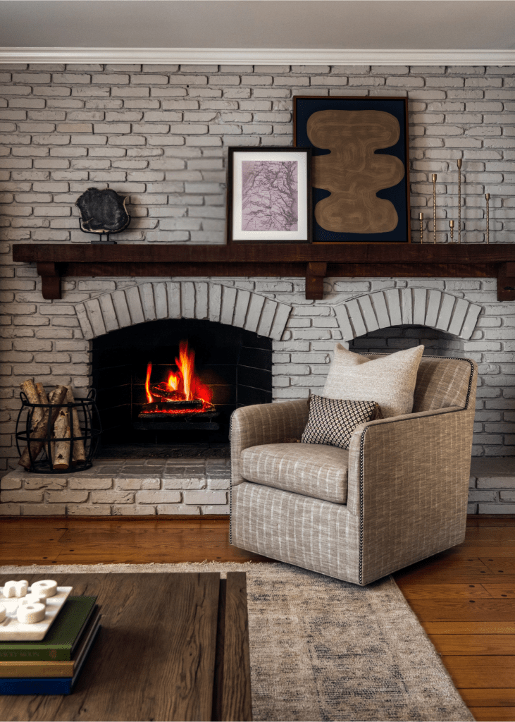 Cozy living room with a gray armchair, a hardwood floor, and a rustic stone fireplace with flames, decorated with wall art and a wooden mantel.