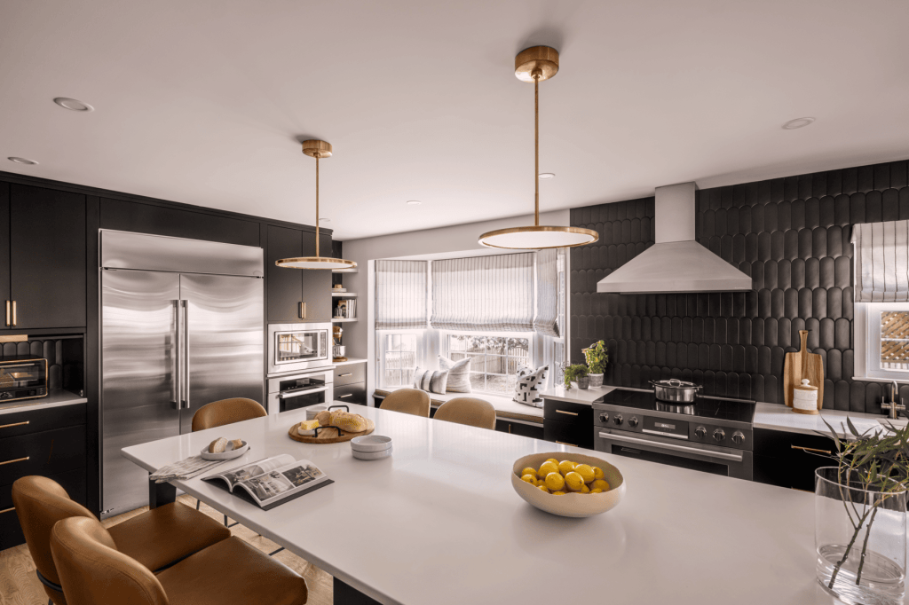 Modern kitchen with a large white island, stainless steel appliances, and black cabinetry. Decorative pendant lights hang over the island, featuring a bowl of lemons and a stack of magazines on the countertop.