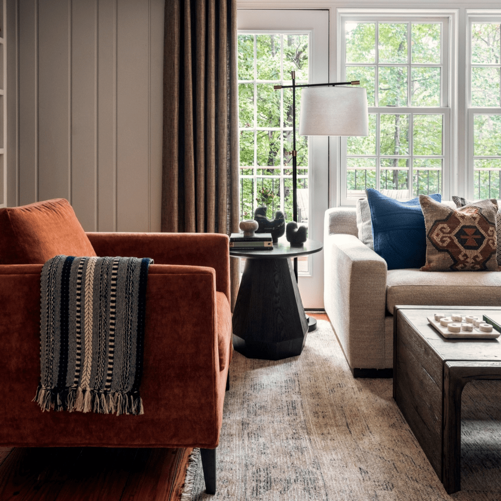 A cozy living room featuring an orange armchair with a patterned throw, a light gray sofa with decorative pillows, a dark side table, a wooden coffee table with decorative items, and large windows allowing natural light to illuminate the space.