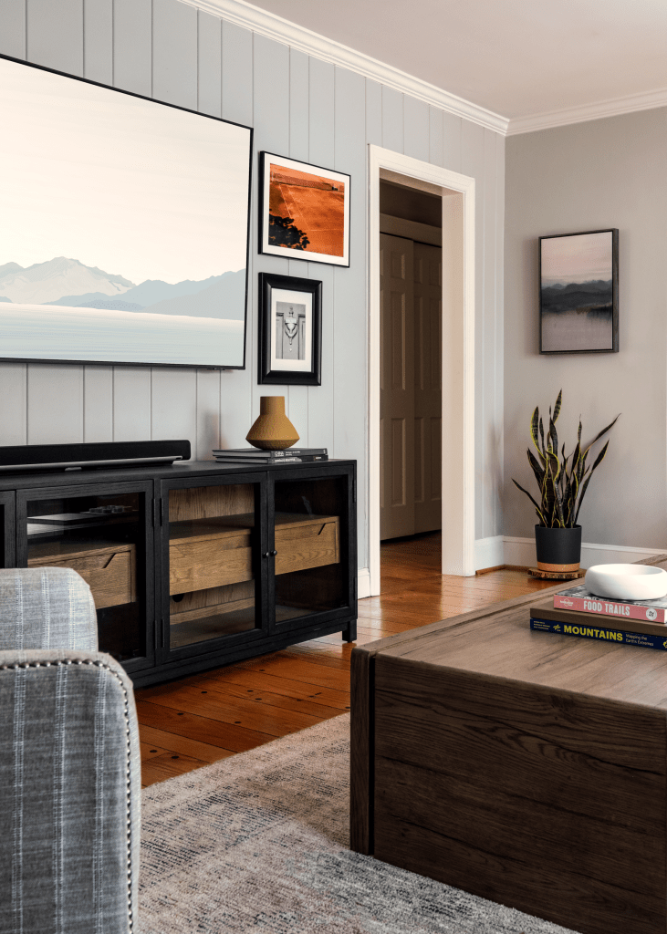 A modern living room featuring a wooden coffee table, a grey upholstered sofa, and a black media console. The wall is adorned with framed art pieces, and a large television is mounted above the console. A doorway leads to another room, and decorative elements like a vase with branches and books are visible.
